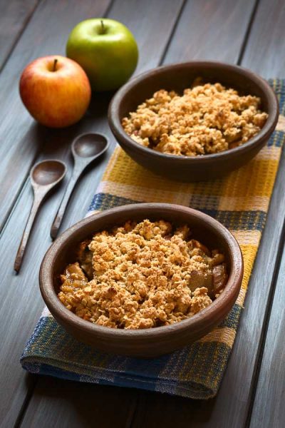 Two rustic bowls of microwave apple crumble on a kitchen towel, wooden spoons and fresh apples on the side, photographed on dark wood with natural light