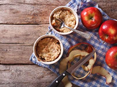 Two cups of freshly made microwave apple crumble, resting on a blue checkered cloth with apples. Horizontal view from above, rustic style