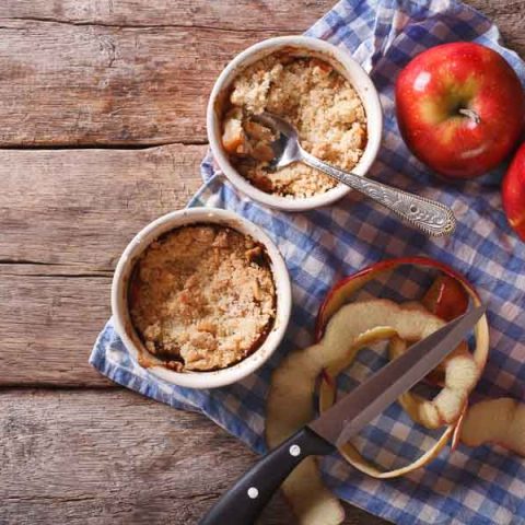 Two cups of freshly made microwave apple crumble, resting on a blue checkered cloth with apples. Horizontal view from above, rustic style