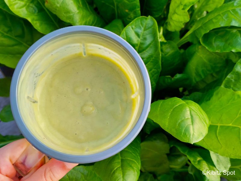 An avocado banana smoothie recipe in a clear cup with a grey lid, held over a patch of growing spinach leaves
