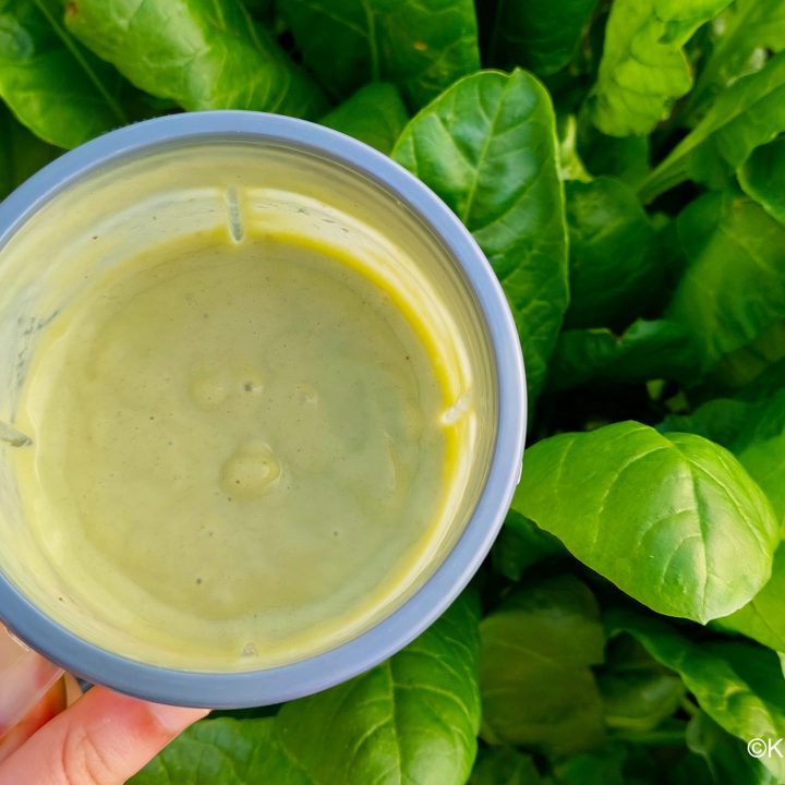 An avocado banana smoothie recipe in a clear cup with a grey lid, held over a patch of growing spinach leaves