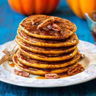 A stack of butternut squash pancakes on a white plate with a blue tablecloth, topped with syrup and pecan nuts
