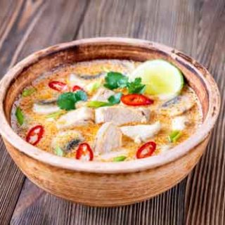 A bowl of Tom Kha Gai on a wooden background. The bowl is full of a creamy orange soup, with chunks of spicy red peppers, chicken, and a slice of lime