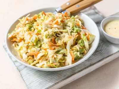 A bowl of our dairy free coleslaw ready to eat. It rests on a wooden block with a side of white sauce