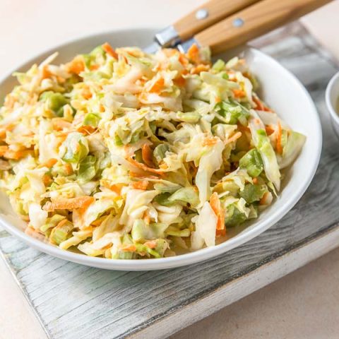 A bowl of our dairy free coleslaw ready to eat. It rests on a wooden block with a side of white sauce