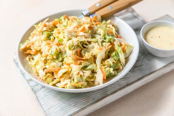 A bowl of our dairy free coleslaw ready to eat. It rests on a wooden block with a side of white sauce