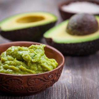 Creamy Guacamole Recipe in a bowl on a wooden table, with a cut up avocado in the background