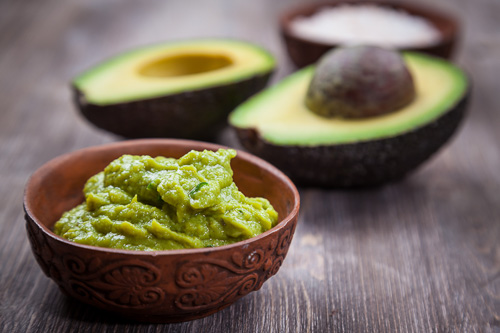 Creamy Guacamole Recipe in a bowl on a wooden table, with a cut up avocado in the background