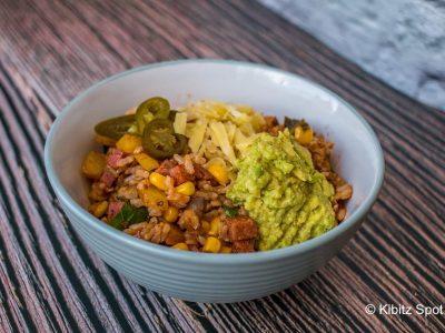 A bowl of Mexican fried rice on a wooden background. Rice with orange spices and chopped vegetables, garnished with jalapeños, guacamole, and grated cheese