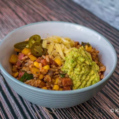 A bowl of Mexican fried rice on a wooden background. Rice with orange spices and chopped vegetables, garnished with jalapeños, guacamole, and grated cheese