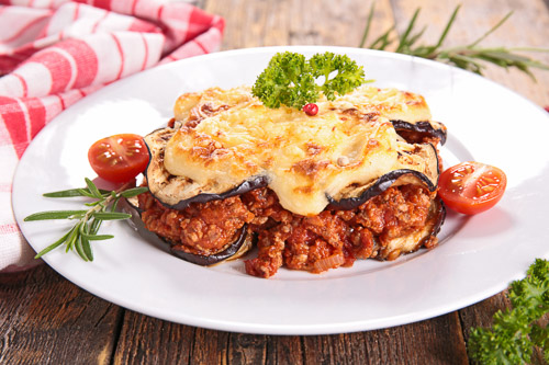 Gluten free moussaka on a white plate with parsley and tomato to garnish. Moussaka has layers of baked eggplant, a red mince sauce, and crispy yellow cheese.