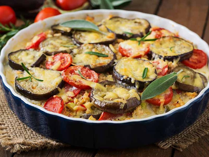 A gluten free moussaka in a black baking pan. It is topped with slices of eggplant, fresh tomatoes, and basil, and slathered in cheesy bechamel sauce