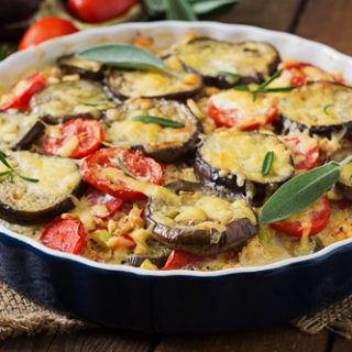 A gluten free moussaka in a black baking pan. It is topped with slices of eggplant, fresh tomatoes, and basil, and slathered in cheesy bechamel sauce