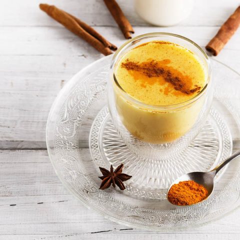 A cup of our turmeric almond milk latte, garnished with cinnamon and plated with spices on a light wooden background.