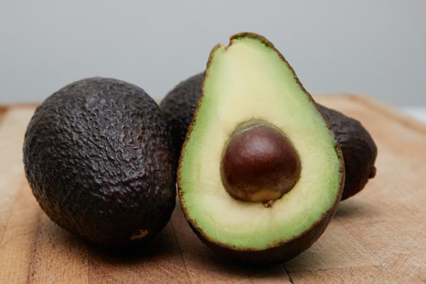 A pile of several whole avocados and one cut avocado on a wooden chopping board. One of the ingredients for an avocado banana smoothie recipe. 