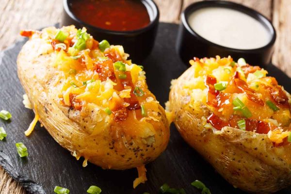Easy Baked Potato Bar Ideas That Meet Everyone's Dietary Requirements 2 Baked potatoes topped with cheddar cheese, green onions and bacon close-up on the table. horizontal