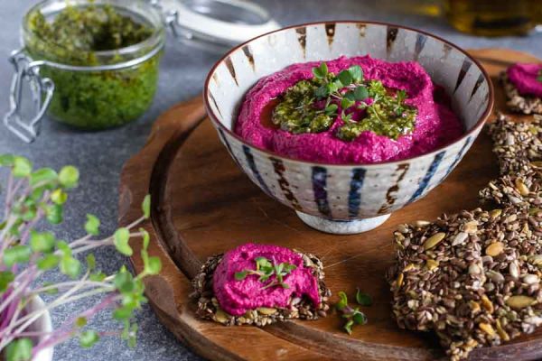A bowl of beetroot hummus on a wooden serving platter with gluten-free seeded crackers