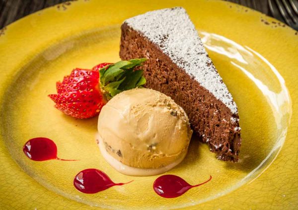 Slice of flourless chocolate cake with salted caramel ice cream siiting on yellow plate with strawberry slices and sauce