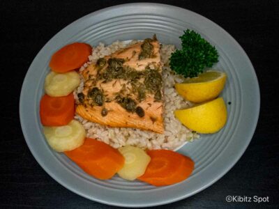 Plate of baked salmon with capers on a bed of brown rice with carrots, parsnips and lemon wedges.