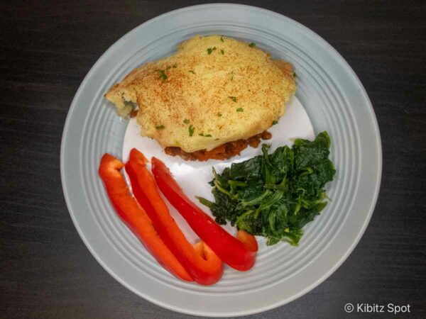 A plate of vegan lentil shepherd's pie with bell pepper slices, and steamed spinach.